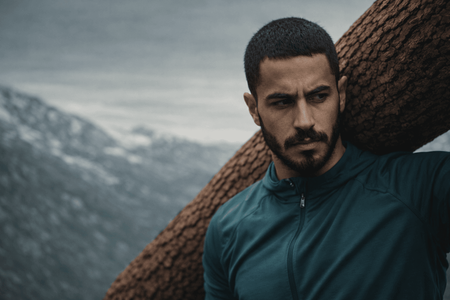 Joe Hachem carrying a heavy tree log over his shoulder in a mountainous landscape, wearing a teal athletic jacket, with a focused and determined expression